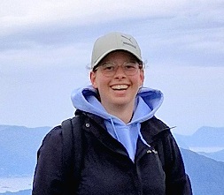 person with a beige cap and glasses smiling with open mouth wearing a purple hoodie with a open black jacket over